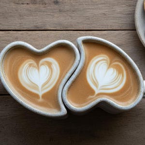 May include: Two heart-shaped mugs filled with latte art, each featuring a white heart design on a creamy brown surface. The mugs are light gray with speckled details and sit on a wooden surface. A plate of cookies is in the background.