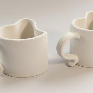 May include: Two off-white ceramic mugs with heart-shaped rims and curved handles. The mugs have a textured surface and are placed side by side on a white background. The handles curl at the base.