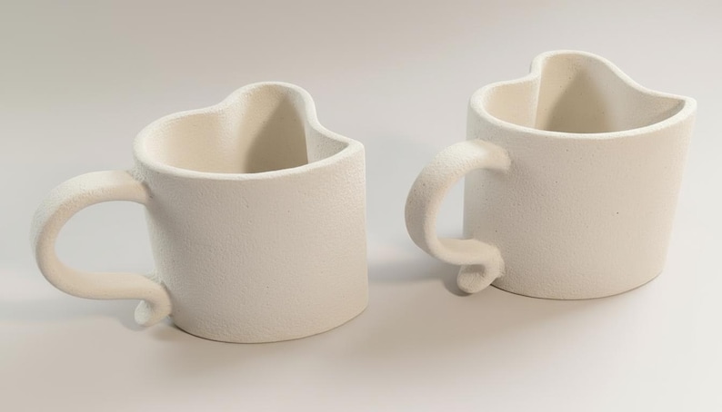 May include: Two off-white ceramic mugs with heart-shaped rims and curved handles. The mugs have a textured surface and are placed side by side on a white background. The handles curl at the base.