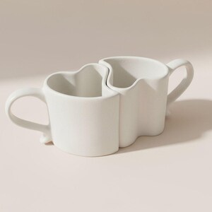 May include: Two white ceramic mugs interlock to form a unique shape. Each mug has a curved handle and a wavy top edge. The mugs are designed to fit together, creating a cohesive and modern look. Ideal for coffee or tea.