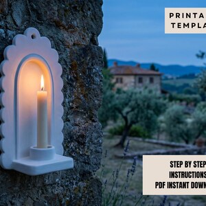 a white candle holder mounted on a stone wall, with a lit candle inside. In the background, there is a house surrounded by trees, creating a serene and peaceful atmosphere.