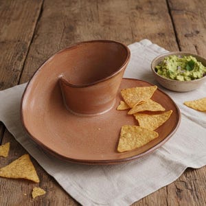 A brown ceramic plate with a unique, spiral-shaped design, containing tortilla chips and a small bowl of guacamole.