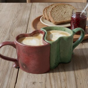May include: Two ceramic mugs, one burgundy and one green, are nestled together, each filled with coffee. A wooden tray holds bread slices and a jar of jam. The mugs have heart-shaped tops and are on a wooden table.