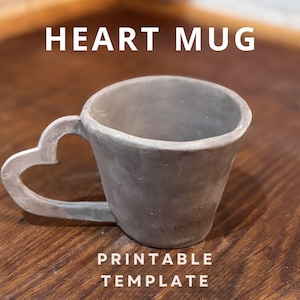 May include: A gray ceramic mug with a heart-shaped handle. The mug is on a wooden surface. The text "HEART MUG PRINTABLE TEMPLATE" is below the mug.