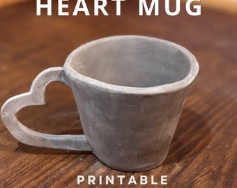 May include: A gray ceramic mug with a heart-shaped handle. The mug is on a wooden surface. The text "HEART MUG PRINTABLE TEMPLATE" is below the mug.