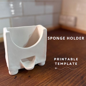 Peut inclure: Porte-éponge en céramique blanche avec un dessus incurvé et quatre petits pieds. Le porte-éponge est conçu pour contenir une éponge et drainer l'excès d'eau. Le texte "SPONGE HOLDER PRINTABLE TEMPLATE" est visible sur l'image.