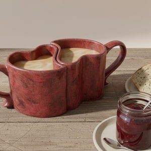 May include: A pair of heart-shaped red mugs filled with coffee, joined together. Beside the mugs are slices of bread on a plate, and a jar of jam with a spoon. The mugs have a marbled texture.