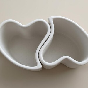 May include: Two white ceramic mugs, each shaped like half a heart, interlock to form a complete heart shape. Each mug has a handle. The mugs are on a neutral background.
