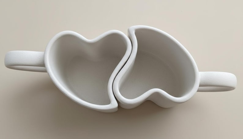 May include: Two white ceramic mugs, each shaped like half a heart, interlock to form a complete heart shape. Each mug has a handle. The mugs are on a neutral background.