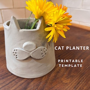 May include: A grey ceramic cat planter with a cute face, holding a bouquet of yellow flowers. The planter has the text "CAT PLANTER PRINTABLE TEMPLATE" on the background.
