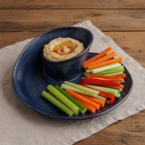 A blue ceramic plate with a curved design holds a serving of hummus and a variety of colorful vegetables, including carrots and celery, arranged on a white cloth.