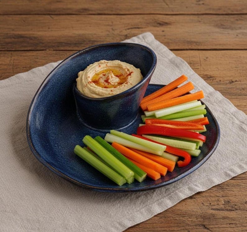 A blue ceramic plate with a curved design holds a serving of hummus and a variety of colorful vegetables, including carrots and celery, arranged on a white cloth.