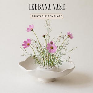 May include: A white ceramic Ikebana vase with a wide, scalloped rim, holding pink cosmos flowers and greenery. The vase has a round base and a kenzan flower frog. Text on the image reads "IKEBANA VASE" and "PRINTABLE TEMPLATE."