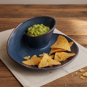 A blue ceramic bowl filled with guacamole dip is placed on a wooden table, accompanied by a plate of tortilla chips.