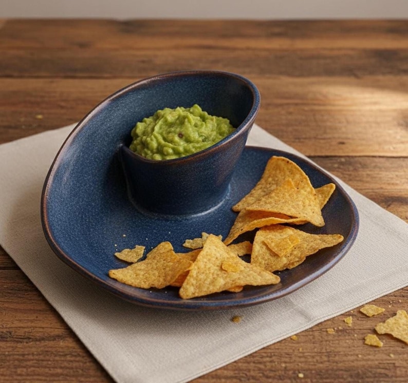 A blue ceramic bowl filled with guacamole dip is placed on a wooden table, accompanied by a plate of tortilla chips.