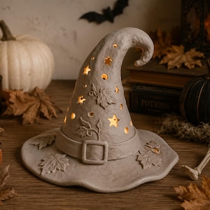 A white witch's hat with carved leaves and stars sits on a wooden surface, surrounded by Halloween decorations such as a skeleton, pumpkins, and candles.