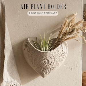 May include: A heart-shaped wall-mounted air plant holder with a textured, carved surface. The holder contains a green air plant and dried wheat stalks. Text at the top reads AIR PLANT HOLDER and PRINTABLE TEMPLATE in black and dark brown.