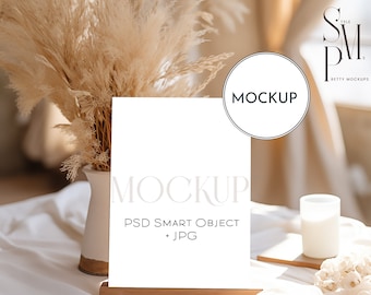 Puede incluir: Una maqueta de tarjeta blanca con el texto "MOCKUP PSD SMART OBJECT + JPG" en un caballete de madera. La tarjeta está sobre una superficie blanca con un jarrón de hierba de la pampa seca y una vela.
