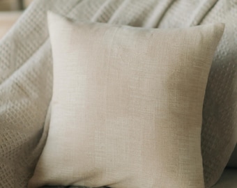 May include: A square, beige linen pillow with a textured surface. The pillow is placed on a light beige, waffle-weave blanket. The pillow has a simple, minimalist design, suitable for home decor.