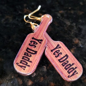 May include: A pair of novelty earrings featuring a paddle design with the text "Yes Daddy" in black. The earrings have a wood-grain appearance and gold-colored ear wires.