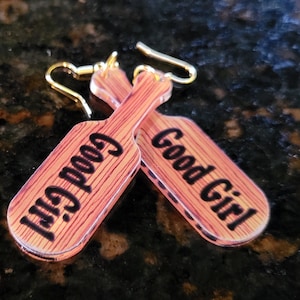 May include: A pair of novelty earrings shaped like paddles. The earrings have a wood grain design with the words "Good Girl" printed in black. The earring hooks are gold-colored.