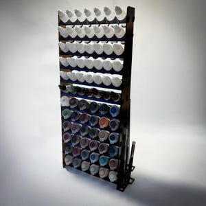 May include: A black paint bottle and brush organiser with multiple tiers. It holds numerous paint bottles with white caps and various coloured paints. Paint brushes are stored on the side. The organiser is designed to hold a large collection of paints.