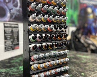 May include: A black wooden paint bottle storage rack with multiple rows of small bottles of paint. The bottles are arranged in a grid pattern and are filled with various colors of paint.