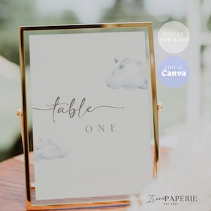 May include: A vertical rectangular sign in a thin gold metal frame featuring a light gray background with watercolor-style clouds and a small heart. The text reads "table ONE" in a combination of cursive and block lettering, centered on the sign.