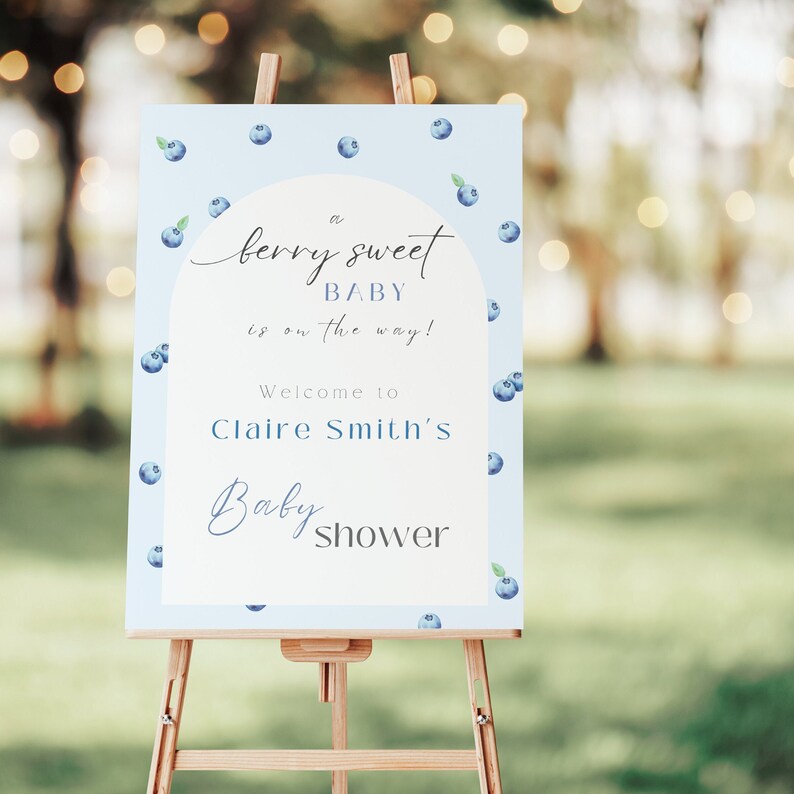 May include: A blue and white baby shower sign with blueberries scattered across the background. The sign reads "a berry sweet baby is on the way! Welcome to Claire Smith's Baby Shower".
