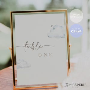 May include: A gold-framed table number card with the word "table" in cursive and "ONE" in block letters. The card features watercolor cloud illustrations and is designed for events. The text "Instant Download" and "Edit in Canva" are also visible.
