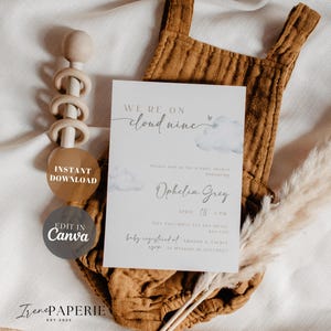 May include: A baby shower invitation with the text "We're on Cloud Nine" and "Ophelia Grey" in elegant script. The invitation features a watercolor cloud design and is set against a brown onesie and a wooden baby rattle.