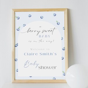 May include: A white sign with a blue and white blueberry pattern. The sign says "a berry sweet baby is on the way!" and "Welcome to Claire Smith's Baby Shower".