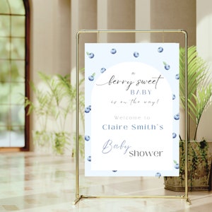 May include: A blue and white baby shower sign with a blueberry theme. The sign reads "A berry sweet baby is on the way! Welcome to Claire Smith's Baby Shower".