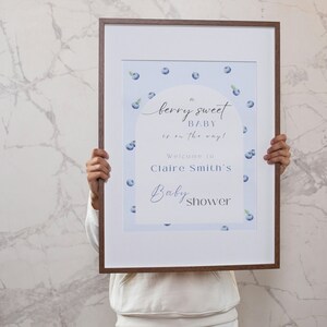 May include: A framed sign with a blue and white blueberry design. The sign reads "A berry sweet baby is on the way! Welcome to Claire Smith's Baby Shower".