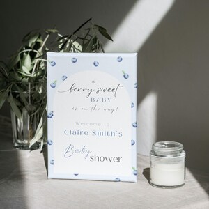 May include: A blue and white baby shower sign with blueberries printed on it. The sign reads "A berry sweet baby is on the way! Welcome to Claire Smith's Baby Shower".