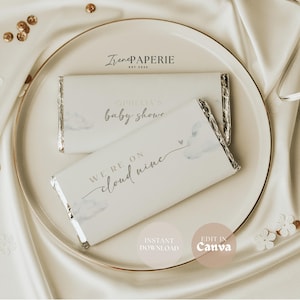 May include: Two white chocolate bars with silver foil wrapping, displayed on a white plate with a gold rim. One bar reads "Ophelia's baby shower," the other says "We're on cloud nine." The background is a white satin cloth.