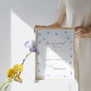 May include: A blue and white baby shower sign with blueberries and the text "A berry sweet baby is on the way! Welcome to Claire Smith's Baby Shower".