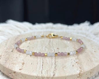 May include: Delicate beaded bracelet featuring alternating pink and white gemstones, accented with small gold-colored beads. The bracelet is secured with a gold-tone clasp. The jewelry is displayed on a light-colored, textured surface.
