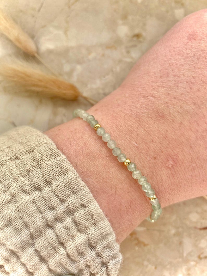 May include: A delicate bracelet featuring small, pale green beads interspersed with gold-colored accents. The bracelet is worn on a wrist, showcasing its subtle elegance. The beads appear to be semi-translucent, adding a touch of sophistication to the jewelry.