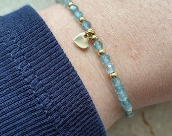 May include: A delicate bracelet made with light blue beads and gold accents. A small gold heart charm hangs from the bracelet.