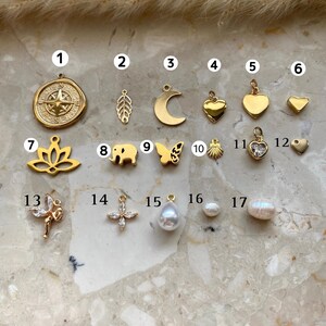 May include: A collection of gold-colored charms and pendants. The charms include a compass rose, leaf, crescent moon, hearts, lotus flower, elephant, butterfly, seashell, and a cross. Also shown are pearl pendants and round pearls.
