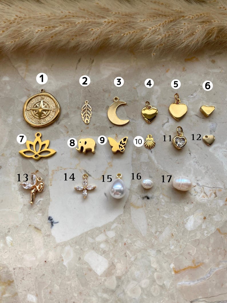 May include: A collection of gold-colored charms and pendants. The charms include a compass rose, leaf, crescent moon, hearts, lotus flower, elephant, butterfly, seashell, and a cross. Also shown are pearl pendants and round pearls.