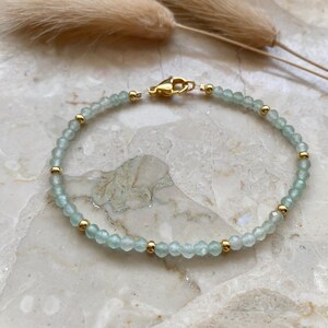 May include: A delicate bracelet featuring small, light blue beads interspersed with gold-colored beads. The bracelet is secured with a gold-colored clasp. The jewelry is displayed on a marble surface, with dried ornamental grass in the background.