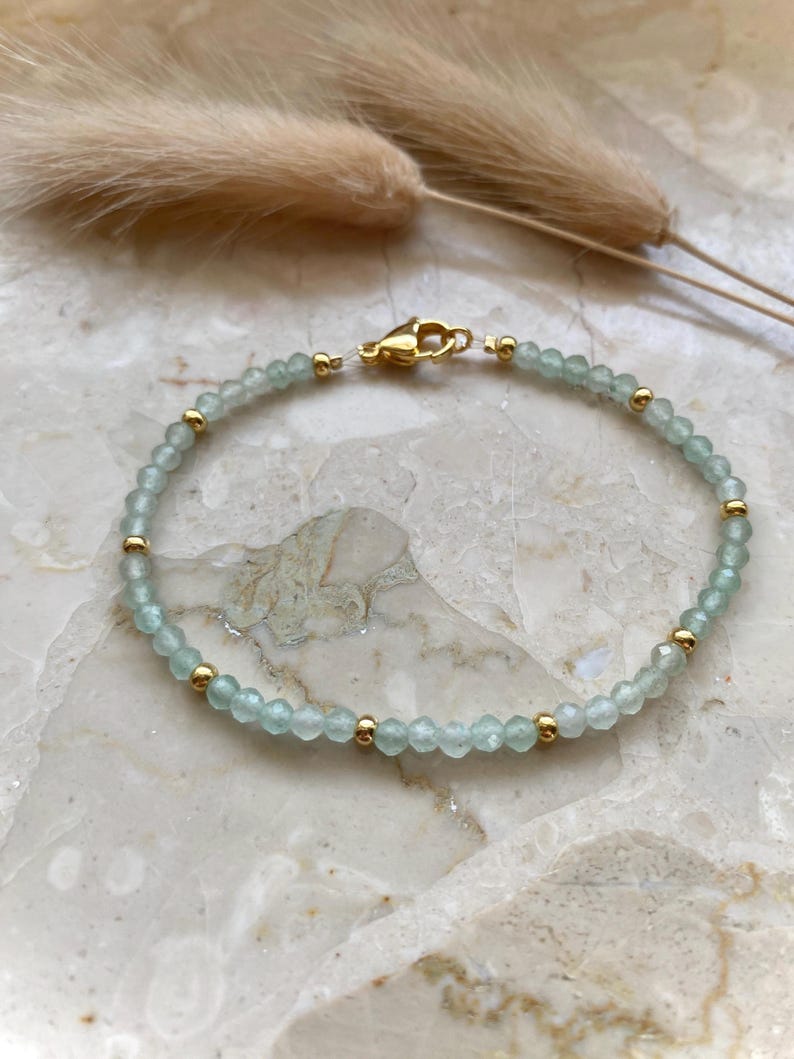 May include: A delicate bracelet featuring small, light blue beads interspersed with gold-colored beads. The bracelet is secured with a gold-colored clasp. The jewelry is displayed on a marble surface, with dried ornamental grass in the background.