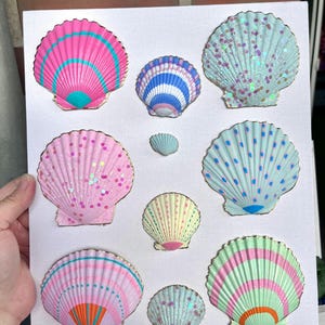 Könnte beinhalten: Eine weiße rechteckige Leinwand mit neun dekorativen Jakobsmuscheln, die in einem Raster angeordnet sind. Die Muscheln sind in verschiedenen Pastellfarben wie Rosa, Blau, Gelb und Grün bemalt und mit goldenen Rändern sowie Glitzerakzenten versehen.