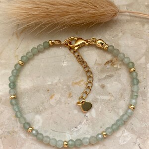 May include: A delicate bracelet featuring pale green, faceted beads interspersed with small gold-colored beads. The adjustable gold-toned chain has a heart-shaped charm. The bracelet is displayed on a marble surface.
