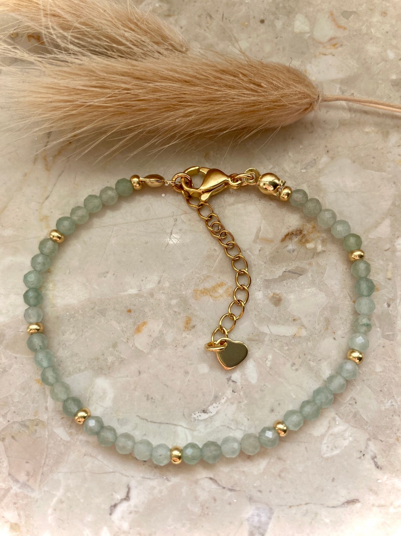 May include: A delicate bracelet featuring pale green, faceted beads interspersed with small gold-colored beads. The adjustable gold-toned chain has a heart-shaped charm. The bracelet is displayed on a marble surface.