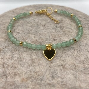 May include: A delicate bracelet featuring small, light green beads and gold-colored accents. A gold heart charm hangs from the center. The bracelet is displayed on a neutral-colored surface.