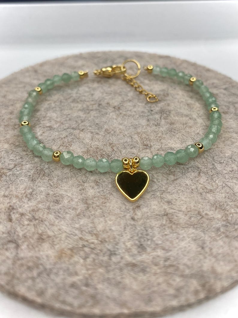 May include: A delicate bracelet featuring small, light green beads and gold-colored accents. A gold heart charm hangs from the center. The bracelet is displayed on a neutral-colored surface.