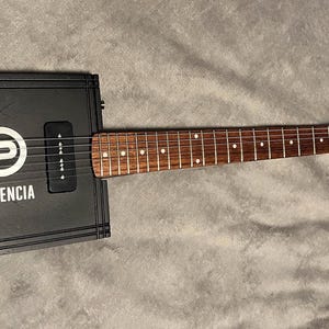 May include: A black electric guitar with a wooden fretboard and a rectangular body. The body has a white "P" logo and the text "PLASENCIA" on it. The guitar has two knobs on the body.